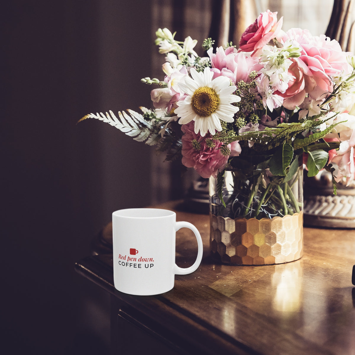 Red Pen Down Coffee Up mug on a dark wood table beside a vase of flowers